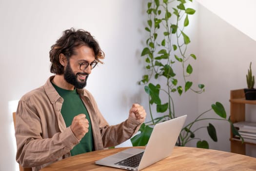 Cheerful man celebrates a success at work with his fists. Happy man reads good news in the laptop. High quality photo