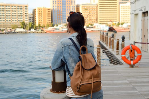 A young woman with a backpack on her back admires the river in the old Dubai Creek. Back view. Journey through the Persian Gulf.