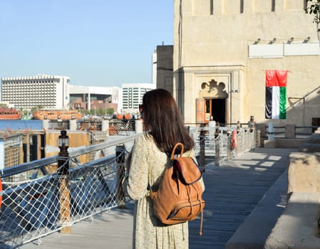 A tourist woman in a beige long dress with a backpack walks along the old narrow streets of Bur Dubai and Creek. Travel and sightseeing concept.