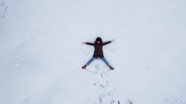 A young woman lies on the snow and makes the figure of an angel. View of the girl from above. The camera moves away. 4k