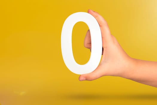 Numeral zero in hand. A hand holds a white number zero on a yellow background with copy space. Zero concept, 0 percent interest rate, minimum air emissions, cost or credit no increase