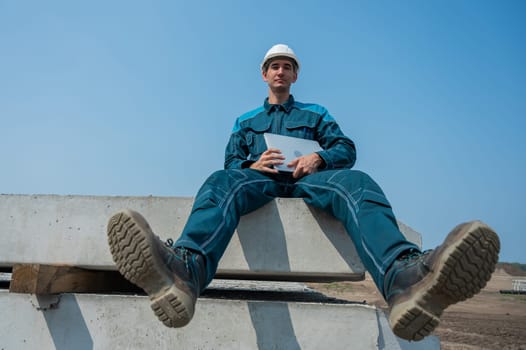 Caucasian male builder in hardhat sits on floor slabs and uses laptop at construction site