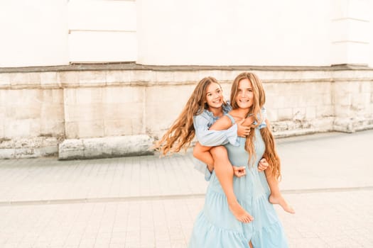 Mother of the daughter walks playing. Mother holds the girl on her back, holding her legs, and her daughter hugs her by the shoulders. Dressed in blue dresses