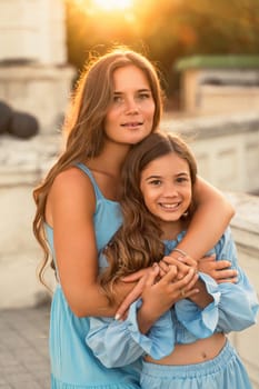 Portrait of mother and daughter in blue dresses with flowing long hair against the backdrop of sunset. The woman hugs and presses the girl to her. They are looking at the camera