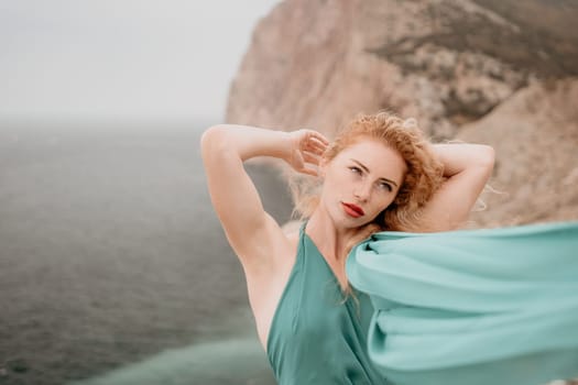 Side view a Young beautiful sensual woman in a mint long dress posing on a volcanic rock high above the sea during sunset. Girl on the nature on overcast sky background. Fashion photo