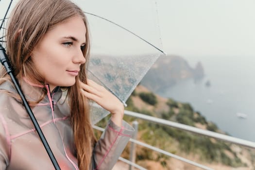 Woman rain park. Happy woman portrait wearing a raincoat with transparent umbrella outdoors on rainy day in park near sea. Girl on the nature on rainy overcast day