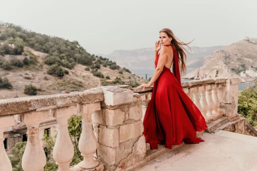 Side view a Young beautiful sensual woman in a red long dress posing on a volcanic rock high above the sea during sunset. Girl on the nature on overcast sky background. Fashion photo