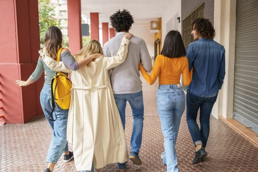 Back view of group of multiethnic friends hugging and walking along street while enjoying stroll in city