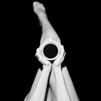 Top view of crop unrecognizable barefooted female sitting in black studio with cup of hot coffee