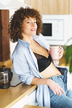 Adult female in unbuttoned shirt and unzipped blue jeans leaning on wooden counter while having cup of hot beverage in modern kitchen