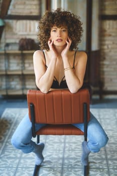 Full body of sensual young female, with afro hairstyle in black lingerie and blue jeans witting on leather chair and touching face while looking at camera against brick wall