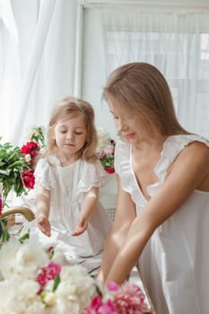 A little blonde girl with her mom on a kitchen countertop decorated with peonies. The concept of the relationship between mother and daughter. Spring atmosphere.