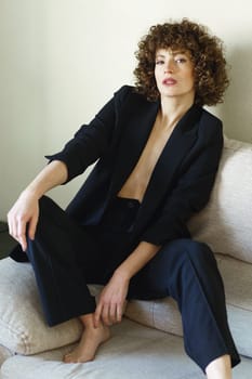 Full body high angle of self-confident female with curly dark hair sitting on couch wearing black jacket and pants looking at camera