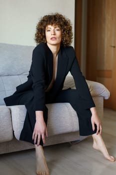 Full body of adult female with dark curly hair wearing black suit sitting on gray sofa leaning forward and looking at camera
