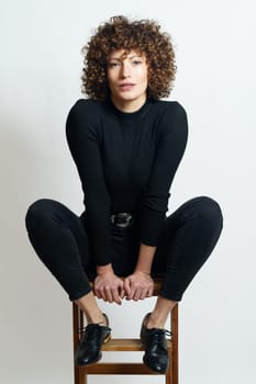Full body of young confident female, model in black outfit relaxing on stool with steps while placing hands between legs on seat and looking at camera against gray backdrop