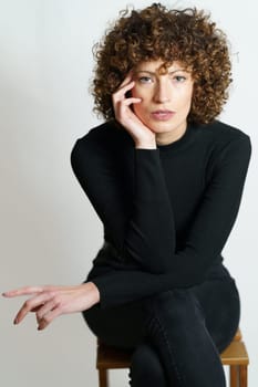 Concentrated and confident young female, in black outfit with hand touching cheek sitting on chair with legs crossed while leaning on elbow and looking at camera in gray backdrop