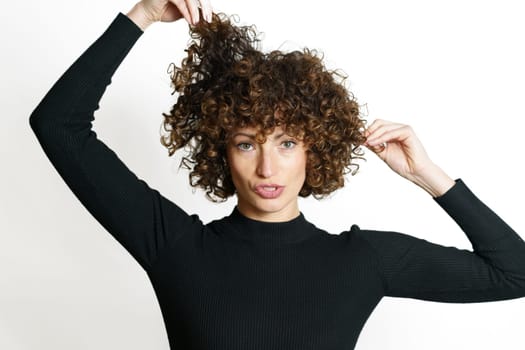 Attractive young female model, with opened mouth and in black sweater looking at camera while pulling curly hair up and down with both hands against gray backdrop