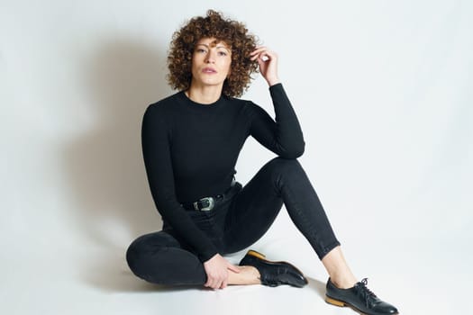 Full body of confident young female model, in trendy outfit relaxing on floor and looking at camera while touching hair with fingers and leaning on raised leg against gray background