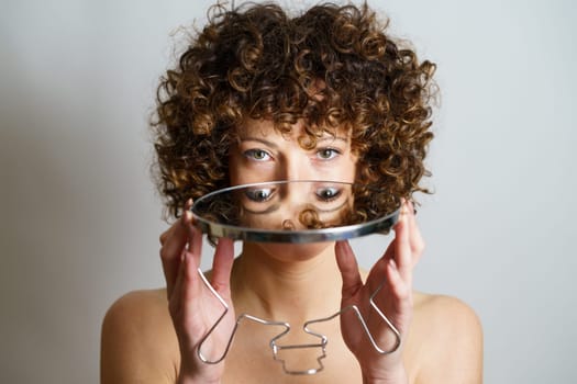 Attractive young curly haired woman model holding round mirror in hands with reflection of face and eyes while looking at camera against gray background