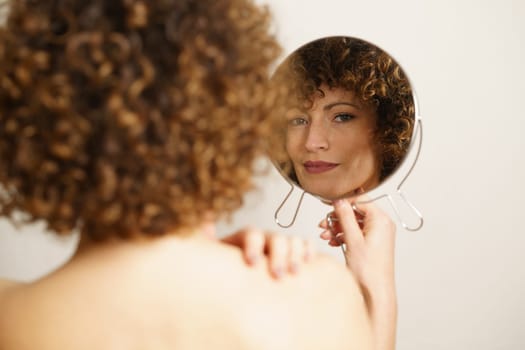 Beautiful young female model, with curly hair and makeup standing with back against camera while holding mirror in hand and looking at camera in reflection near gray background