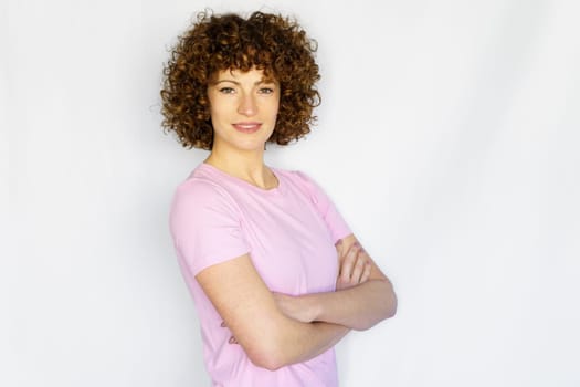 Confident adult curly haired girl model wearing basic pink t-shirt, standing with arms crossed against white background smiling at camera