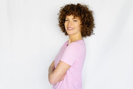 Positive adult girl in casual apparel keeping arms crossed while standing against white background and looking at camera smiling