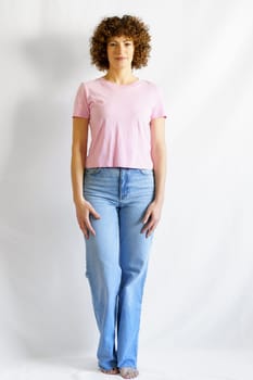 Full body of curly haired female in jeans and pink t-shirt leaning on white wall looking at camera