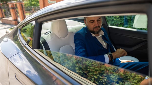 A caucasian man in a blue suit looks at his watch while sitting in the back seat of a car. Business class passenger