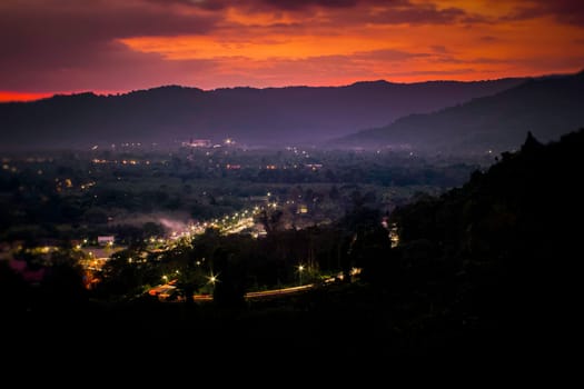 Landscape with alpine mountain valley, low clouds, forest, purple sky with stars, city illumination at sunset. Aerial.