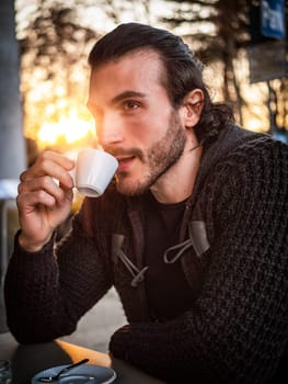 One handsome young man drinking espresso coffee, sitting outside in urban setting in European city