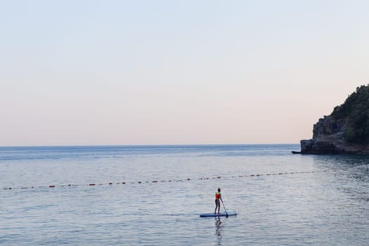 A girl with a paddle in the sea stands on a sapboard at sunrise. Active rest on the sea