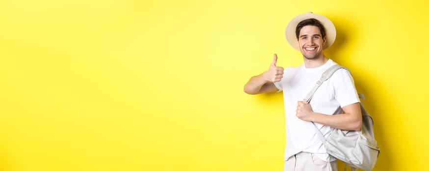 Tourism, travelling and holidays concept. Happy man going on vacation, holding backpack and showing thumb up, smiling satisfied, standing against yellow background.