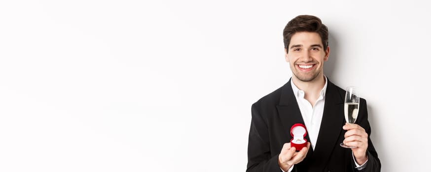 Close-up of handsome man in suit, making a proposal, giving engagement ring and raising glass of champagne, standing against white background.