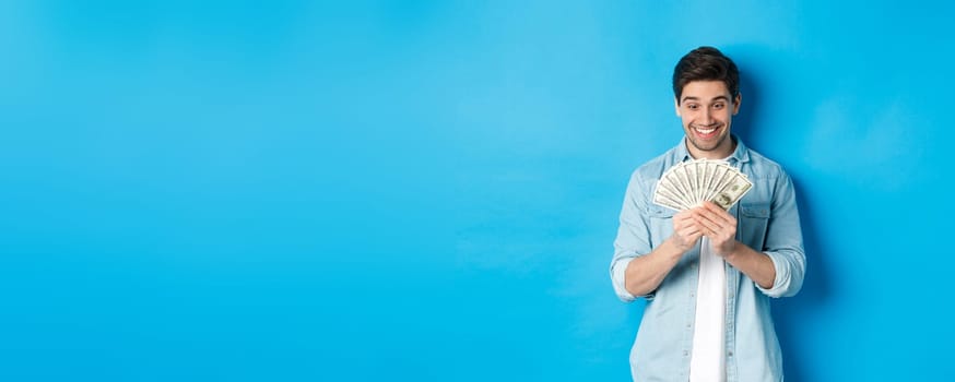 Excited successful man counting money, looking satisfied at cash and smiling, standing over blue background.