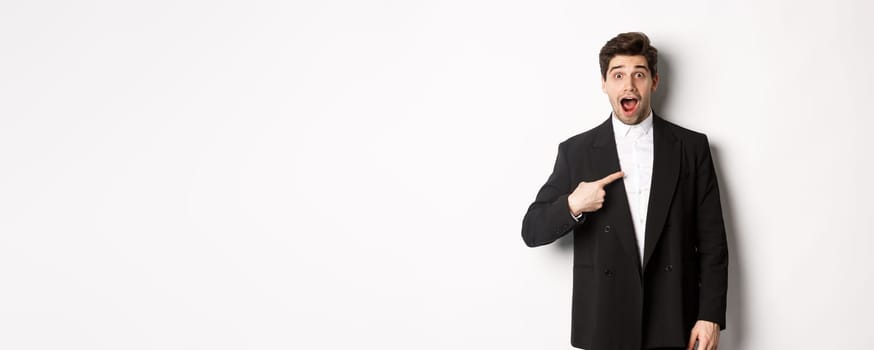 Concept of new year party, celebration and lifestyle. Portrait of surprised handsome guy in black suit, pointing at himself and looking amazed, being chosen, standing over white background.