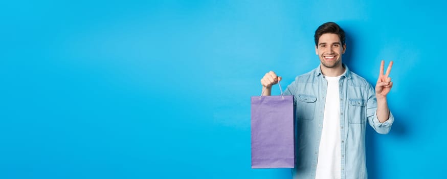Concept of shopping, holidays and lifestyle. Happy bearded guy holding paper bag from store and showing peace sign, standing over blue background.