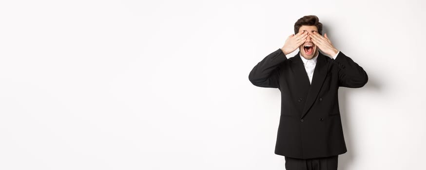 Image of handsome stylish man in black suit, waiting for christmas surprise, covering eyes with hands and smiling, anticipating presents, standing over white background.