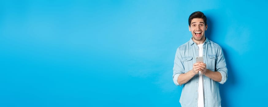 Surprised and happy man winning something online, holding smartphone and rejoicing, standing against blue background.