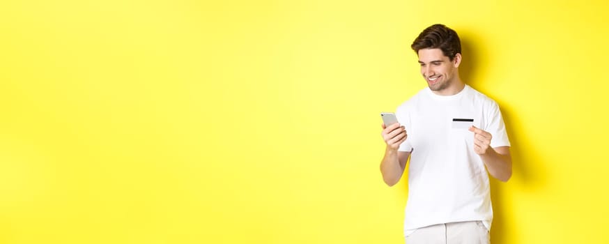 Young man paying online, insert credit card number on mobile phone, shopping in internet, standing over yellow background.