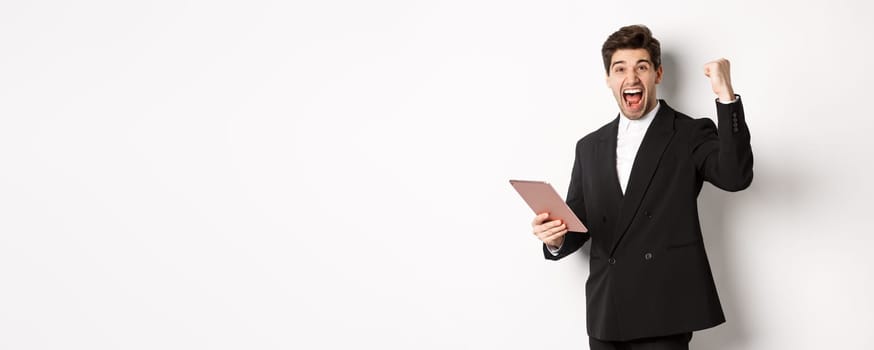 Portrait of handsome successful businessman in black suit, achieve goal, holding digital tablet and saying yes, standing against white background and rejoicing.