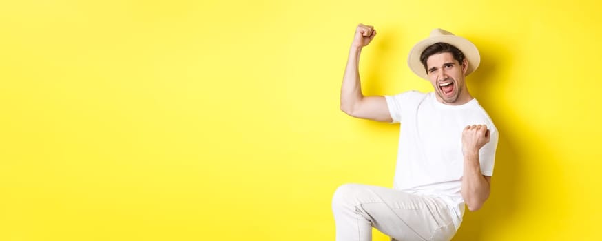 Concept of tourism and vacation. Lucky male tourist winning trip, rejoicing and saying yes, making fist pump while triumphing, standing against yellow background.