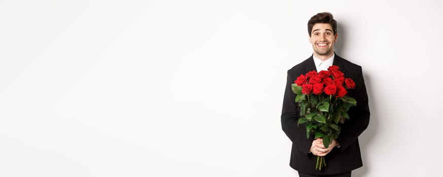 Image of handsome man in black suit, holding bouquet of roses and smiling, standing against white background.