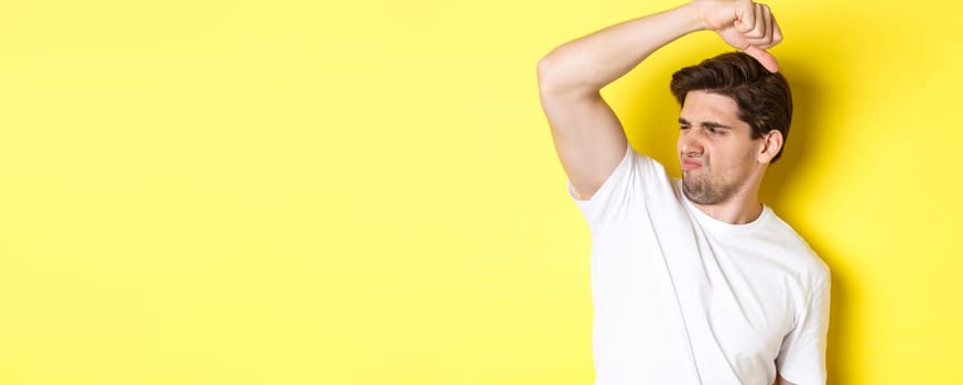 Man in sweat smelling his armpit, standing in white t-shirt and grimacing from stinky clothes.