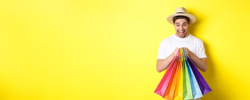 Image of happy man shopping on vacation, holding paper bags and smiling, standing against yellow background.