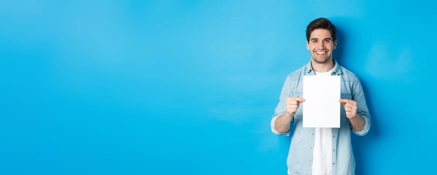 Young smiling guy in casual outfit, holding blank piece of paper with your advertisement, standing over blue background.