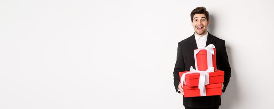 Concept of holidays, relationship and celebration. Handsome man in black suit bringing presents at new year party, holding gifts and smiling amused, standing against white background.