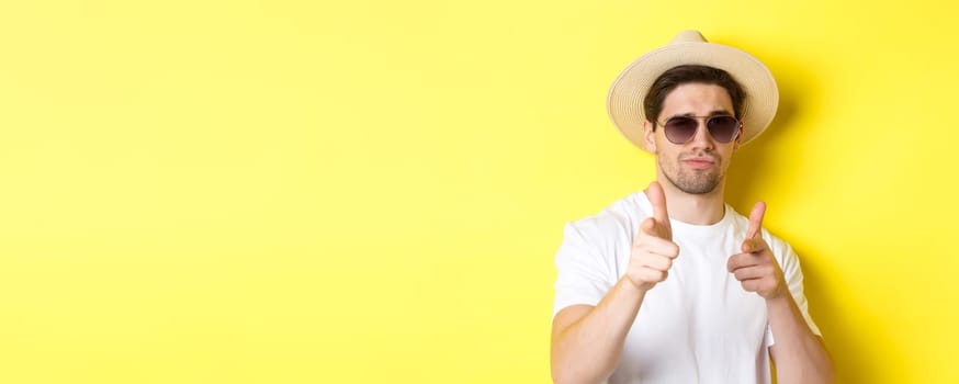 Concept of tourism and vacation. Close-up of cool guy in summer hat and sunglasses pointing finger guns at camera, standing over yellow background.