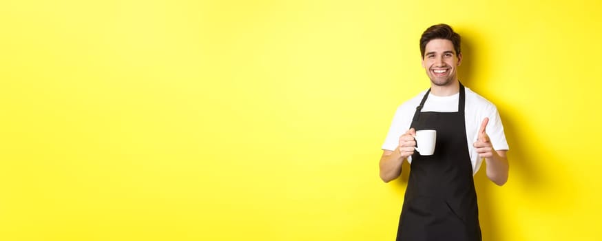 Barista bringing coffee and pointing finger gun at camera, standing in black apron against yellow background.