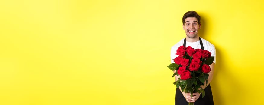 Seller in flower shop wearing black apron, giving bouquet of roses and smiling, standing over yellow background.