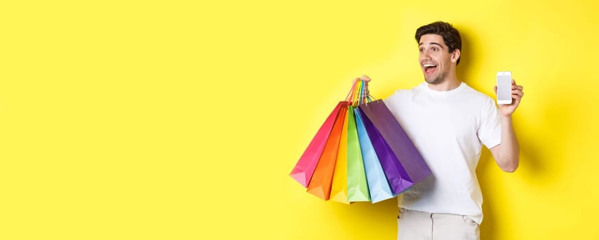 Excited man showing smartphone screen and shopping bags, achieve app goal, demonstrating mobile banking application, yellow background.